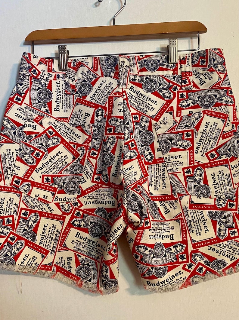 May include: Red and white shorts with a repeating Budweiser label pattern. The shorts are made of a light fabric and have a frayed hem. They are hanging on a wooden hanger.