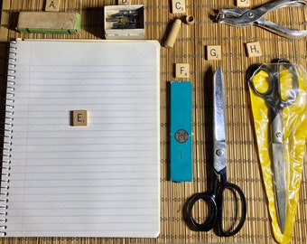 May include: A notebook with lined paper, a tile with the letter E, a box of sewing needles, a pair of scissors, a hole punch, a box of letter tiles, and a pair of scissors in a plastic bag.