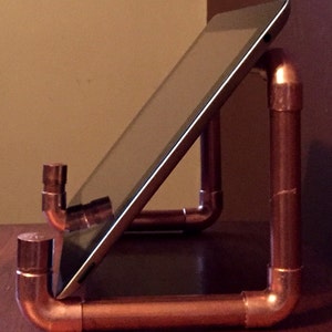 May include: A copper pipe tablet stand with a black tablet propped up at an angle. The stand is made of copper pipes that are connected together with elbows. The stand is on a wooden surface.