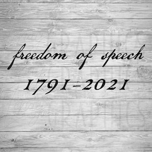 May include: Black text on a gray wooden background reads "freedom of speech" above "1791-2021". The text is in a cursive font. The background is made of horizontal wooden planks.