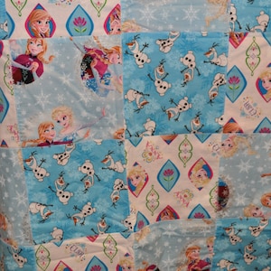 May include: A blue and white patchwork quilt with a Frozen theme. The quilt features images of Elsa, Anna, Olaf, and snowflakes. The quilt is made of various fabrics and is perfect for a child's bedroom.