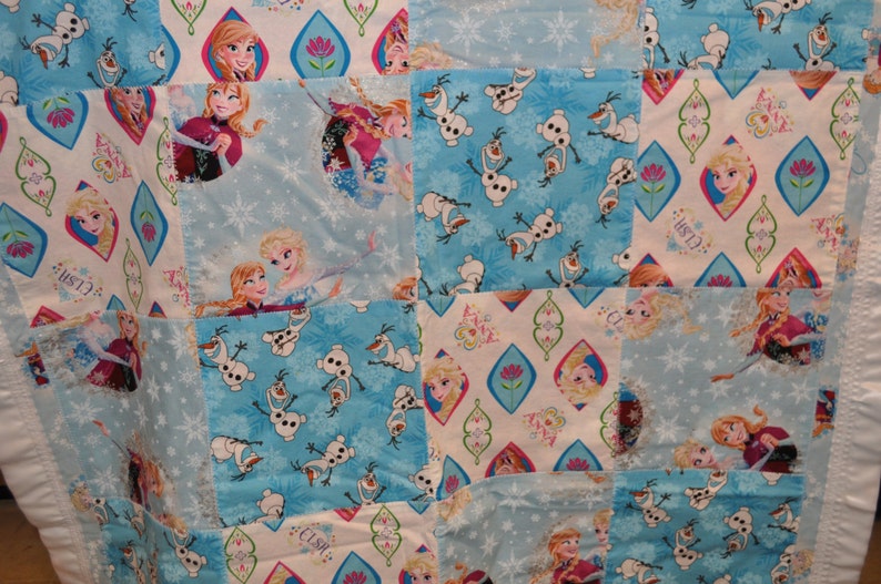 May include: A blue and white patchwork quilt with a Frozen theme. The quilt features images of Elsa, Anna, Olaf, and snowflakes. The quilt is made of various fabrics and is perfect for a child's bedroom.