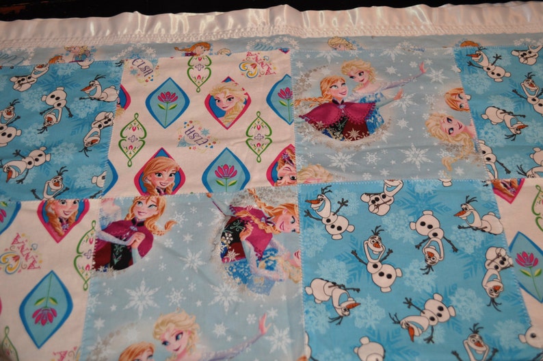 May include: A patchwork quilt featuring a blue and white snowflake pattern, a white background with pink flowers and the words "Elsa" and "Olaf", and a blue background with images of Elsa and Anna from the movie Frozen.