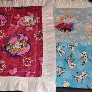 May include: A pink and white baby blanket with a patchwork design featuring images of Elsa and Anna from Frozen and Olaf the snowman. The blanket has a white satin border.