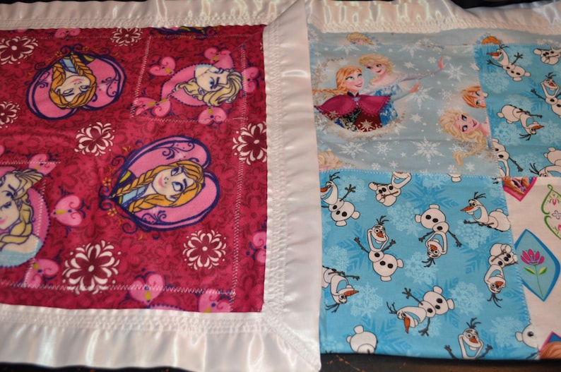 May include: A pink and white baby blanket with a patchwork design featuring images of Elsa and Anna from Frozen and Olaf the snowman. The blanket has a white satin border.