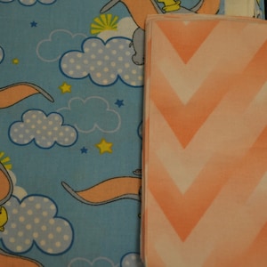 May include: Two fabric swatches, one with a blue background and a repeating pattern of a cartoon elephant flying in the sky with clouds and stars, and the other with a peach background and a chevron pattern.