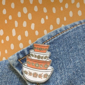 Könnte beinhalten: Eine weiße, orange und grüne Emaille-Anstecknadel mit drei übereinander gestapelten Rührschüsseln mit Blumenmuster.