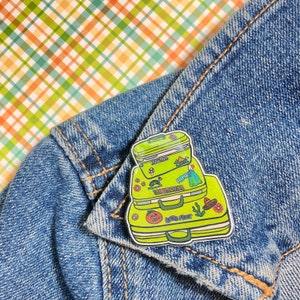 Peut inclure: Une épingle en émail verte et bleue représentant trois valises empilées avec différents motifs et textes sur le thème du voyage, dont "Hawaii" et "Wild Feet".