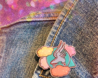 Könnte beinhalten: Eine blaue, rosa und gelbe Emaille-Anstecknadel mit einer Rollschuh mit Flügeln, die durch Wolken fliegen. Die Anstecknadel ist an einer Jeansjacke befestigt.