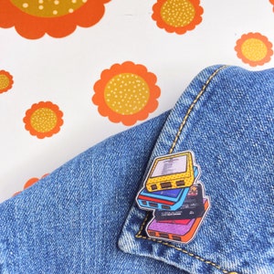 Peut inclure: Une pile de quatre cassettes colorées avec un design jaune, bleu, rouge et violet. Les cassettes sont empilées les unes sur les autres et ont un look rétro. Les cassettes sont épinglées sur une veste en jean bleue.