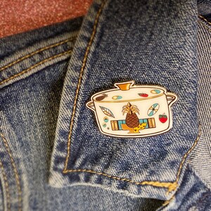 May include: A gold enamel pin depicting a white pot with a pineapple, a strawberry, and other designs. The pin is attached to a blue denim jacket.