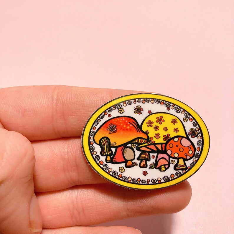 May include: A yellow oval sticker with a white background featuring three cartoon mushrooms with red, orange, and yellow caps. The mushrooms are surrounded by a border of yellow flowers and green leaves.