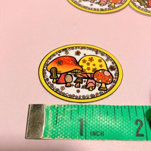 May include: A round sticker with a white background and a yellow border. The sticker features a group of three cartoon mushrooms with red, orange, and yellow caps. The mushrooms are surrounded by a border of small yellow flowers.