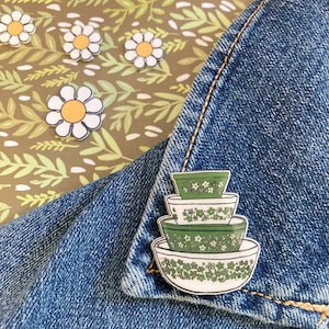 以下が含まれることがあります： 緑と白のエナメル製のミキシングボウルピン。ピンは、花柄の4つの積み重ねられたボウルを特徴としています。ピンは青いデニムの背景にあります。