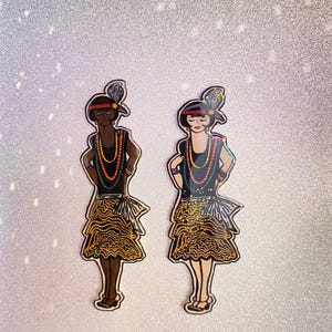 May include: Two flapper-style figures with black dresses, gold beaded necklaces, and gold sequined skirts. One figure has brown skin and the other has light skin. Both figures have black hair and are wearing headbands with feathers.