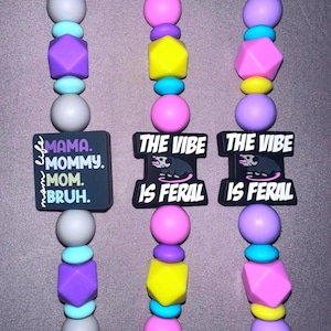 May include: Three beaded lanyards with a variety of colorful beads in pink, purple, yellow, and blue. One lanyard has a black square with the text "MAMA. MOMMY. MOM. BRUH." The other two have a black rectangle with the text "THE VIBE IS FERAL."