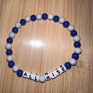 Op de afbeelding: Een blauwe en witte kralen armband met de woorden "ALL RISE" gespeld in witte letter kralen.
