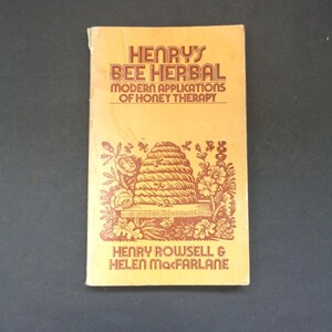 May include: A vintage book titled "Henry's Bee Herbal" with the subtitle "Modern Applications of Honey Therapy." The cover is a light orange color with brown text and illustrations of a beehive and flowers.