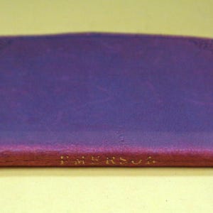 May include: A closed, rectangular book with a purple cover and a red spine. The text "EMERSON" is printed in gold on the spine. A black bookmark extends from the top edge of the book.