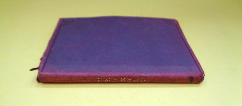 May include: A closed, rectangular book with a purple cover and a red spine. The text "EMERSON" is printed in gold on the spine. A black bookmark extends from the top edge of the book.