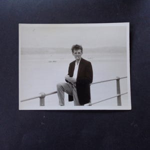 May include: A black and white photo of a man standing on a pier, wearing a dark suit jacket and holding a cigarette. He is looking off to the side.
