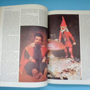 May include: An open book with a red and white illustration of a man in a red and black robe. The book is open to a page with text about the history of little people. The opposite page has a color photograph of a small red gnome standing on a large mushroom.