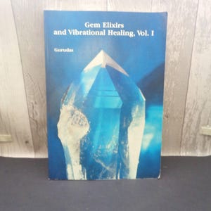 May include: A book titled "Gem Elixirs and Vibrational Healing, Vol. I" by Gurudas. The cover features a large, stylized, blue crystal against a blue background. The book is standing upright.