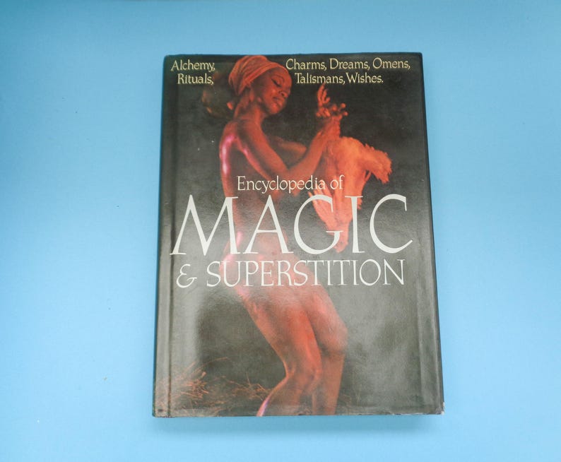May include: A book titled "Encyclopedia of Magic & Superstition" with a black cover and a woman with dark skin and a white head wrap on the front. The book cover also includes the text "Alchemy Rituals, Charms, Dreams, Omens, Talismans, Wishes."