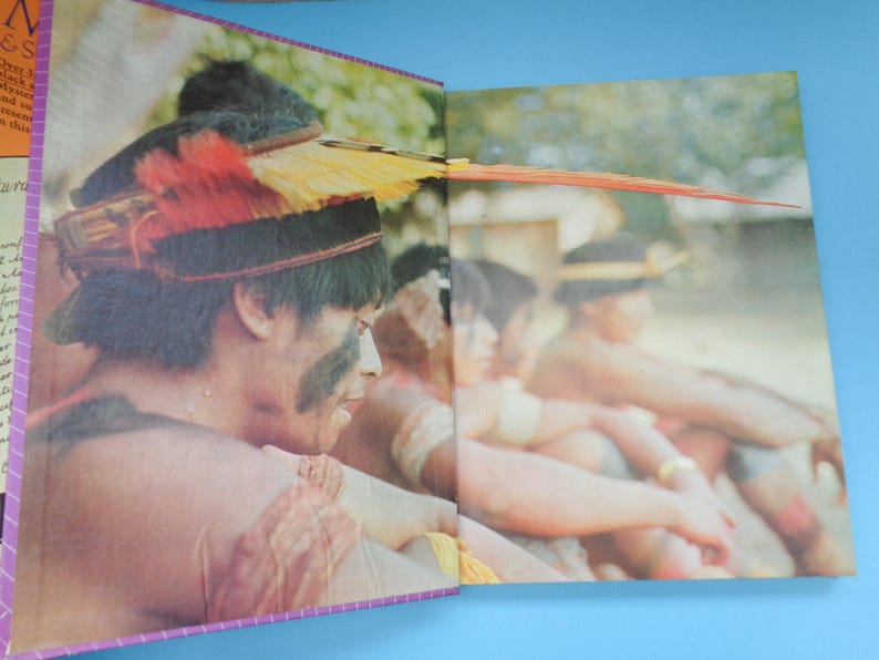 May include: A group of people wearing traditional feathered headdresses and clothing. The people are sitting in a row, and the person in the foreground is looking down.