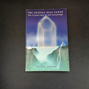 May include: A book titled "THE CRYSTAL ALLY CARDS" with the subtitle "The Crystal Path to Self Knowledge" by Naisha Ahsian. The cover features a large, glowing crystal over a landscape with a blue and green color scheme.