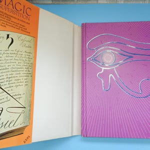 May include: A book titled "Magic & Superstition" with an orange cover and a close-up of the text "Saturday 5 Cassiel" and a drawing of a compass. The back cover is purple with a blue and white drawing of an eye.