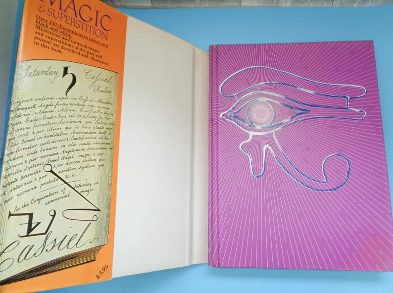 May include: A book titled "Magic & Superstition" with an orange cover and a close-up of the text "Saturday 5 Cassiel" and a drawing of a compass. The back cover is purple with a blue and white drawing of an eye.