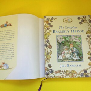 May include: An open book titled "The Complete Brambly Hedge" by Jill Barklem. The cover features an illustration of woodland creatures. The book's pages are cream-colored, and the spine has a purple ribbon bookmark.