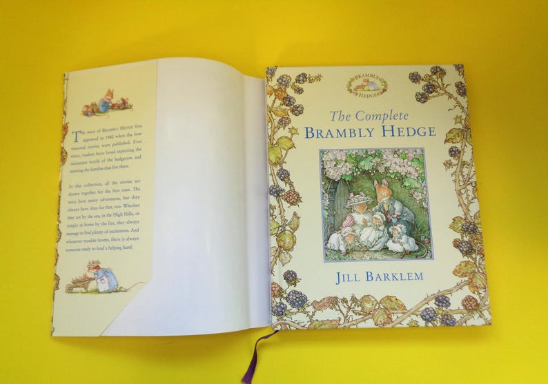 May include: An open book titled "The Complete Brambly Hedge" by Jill Barklem. The cover features an illustration of woodland creatures. The book's pages are cream-colored, and the spine has a purple ribbon bookmark.