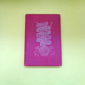 Puede incluir: Un libro vintage de color magenta titulado "Complete Home Knitting Illustrated". La portada presenta texto plateado e ilustraciones de agujas de tejer, hilo y conejos. El libro está cerrado y sobre un fondo amarillo.
