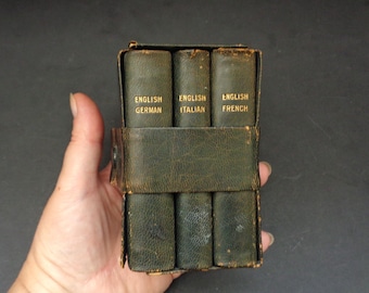 May include: A set of six small, dark green books in a leather case. The books are labeled "English German", "English Italian", and "English French". The case has a strap to hold the books together. The books are held in a hand.