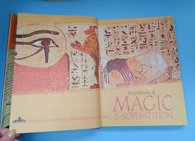 May include: An open book titled "Encyclopedia of Magic & Superstition" with illustrations of an Egyptian eye and a woman lying down. The book cover is yellow with red and black text.