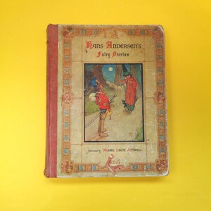 Puede incluir: Libro antiguo de tapa dura titulado "Hans Andersen's Fairy Stories" con ilustraciones de Mabel Lucie Attwell. La portada presenta una colorida ilustración de un soldado y una figura con abrigo rojo. El libro tiene lomo rojo y borde decorativo.