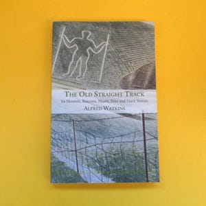 Può includere: Un libro intitolato "The Old Straight Track" di Alfred Watkins. La copertina presenta un disegno di una figura con due linee e un paesaggio con filo spinato. Il libro è su sfondo giallo.