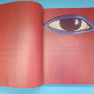 May include: An open book with a red cover and a blue eye illustration on the front. The title of the book is "Encyclopedia of Magic & Superstition".