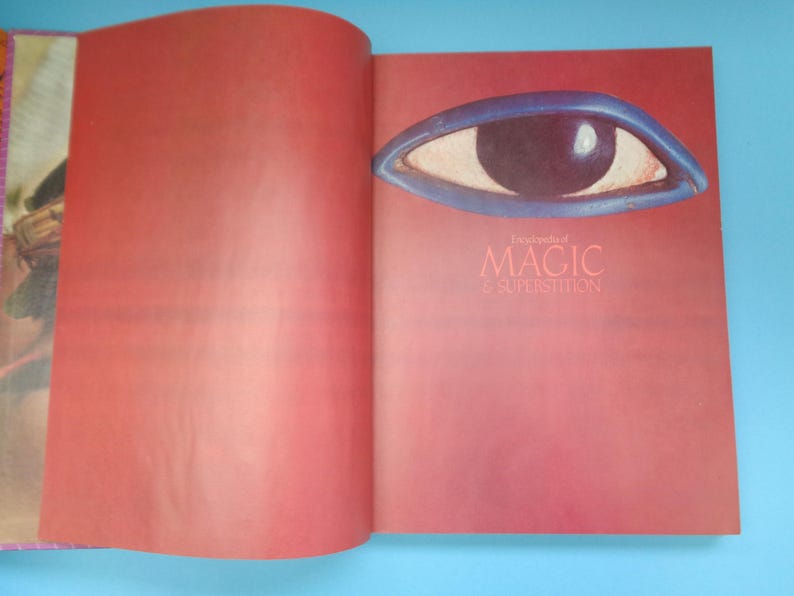 May include: An open book with a red cover and a blue eye illustration on the front. The title of the book is "Encyclopedia of Magic & Superstition".