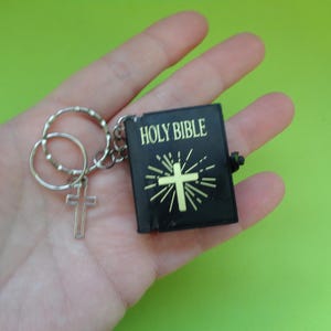 May include: A miniature black keychain featuring a small book titled HOLY BIBLE in gold lettering on the front cover, accompanied by a gold-colored cross symbol with radiating lines and a separate silver-toned metal cross charm attached to the ring.