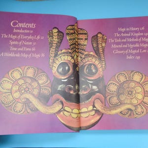 May include: Open book with a purple cover and a colorful illustration of a mask with a large, toothy grin. The book's contents page lists topics such as "The Magic of Everyday Life", "Spirits of Nature", "Time and Form", and "A Worldwide Map of Magic".