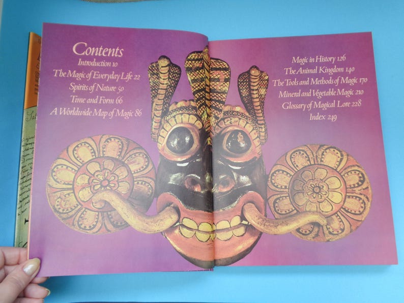 May include: Open book with a purple cover and a colorful illustration of a mask with a large, toothy grin. The book's contents page lists topics such as "The Magic of Everyday Life", "Spirits of Nature", "Time and Form", and "A Worldwide Map of Magic".