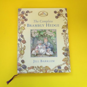 May include: A hardcover book titled "The Complete Brambly Hedge" by Jill Barklem. The cover features an illustration of woodland creatures and is framed by a border of blackberries and leaves. A purple ribbon bookmark is visible.