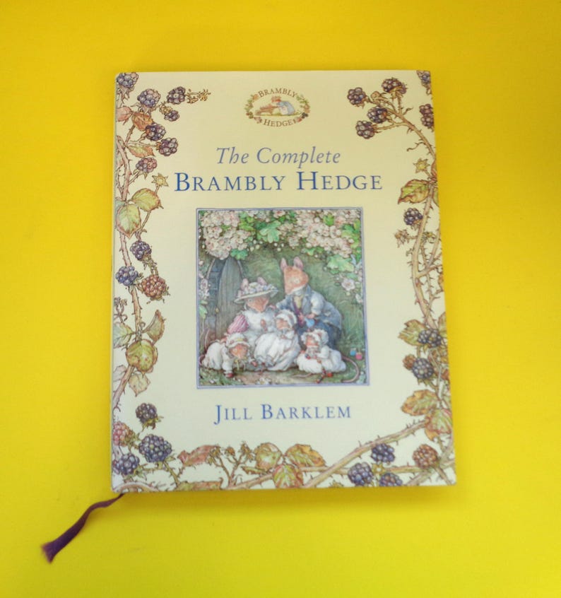 May include: A hardcover book titled "The Complete Brambly Hedge" by Jill Barklem. The cover features an illustration of woodland creatures and is framed by a border of blackberries and leaves. A purple ribbon bookmark is visible.