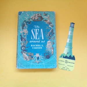 May include: A vintage book titled "The Sea Around Us" by Rachel L. Carson, with a blue cover featuring sea creatures and a ship. A lighthouse-shaped bookmark is next to the book. The bookmark is a Staples Publication.