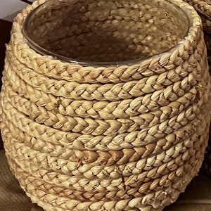 May include: A woven wicker vase with a clear glass insert. The vase is made of natural materials and has a light brown color.