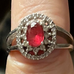 May include: A silver ring with a red oval gemstone surrounded by a halo of smaller clear stones.
