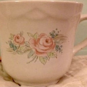 May include: A white ceramic teacup with a floral design featuring pink roses and green leaves. The cup has a rounded shape and a handle.